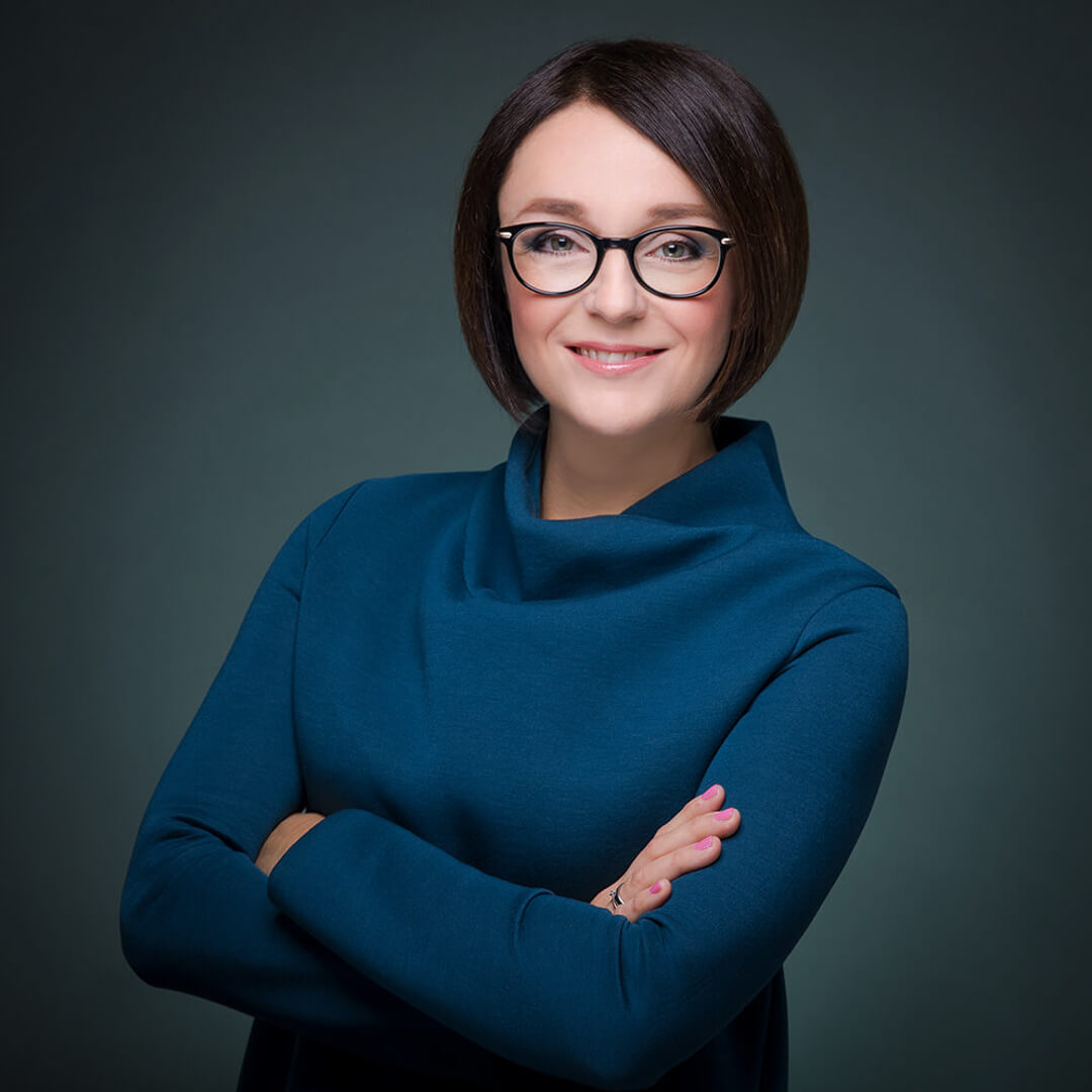 Professionelles Bewerbungsfoto einer Frau mit Brille und blauem Oberteil vor dunklem Studiohintergrund.