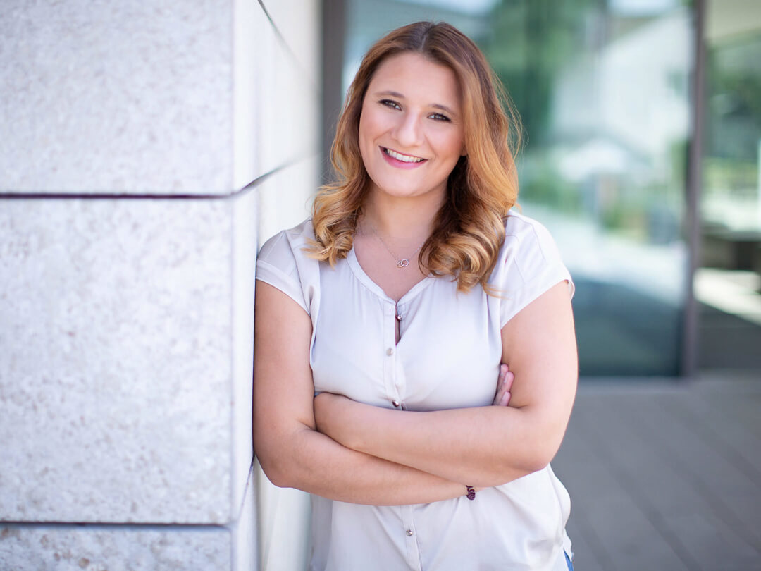 Professionelles Bewerbungsfoto einer lächelnden Frau, die mit verschränkten Armen an einer modernen Steinwand lehnt; aufgenommen im natürlichen Licht vor einem Business-Gebäude.