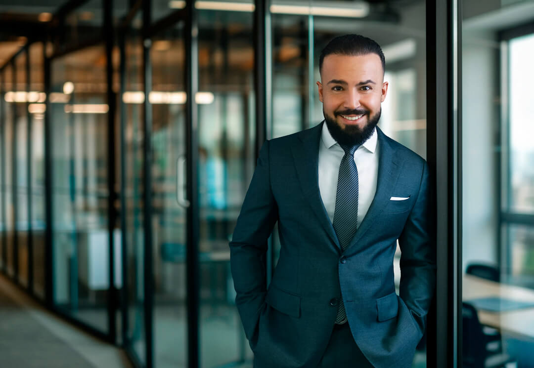 Professionelles Businessfoto eines lächelnden Mannes im dunkelblauen Anzug, aufgenommen in einem modernen Büro mit Glaswänden.